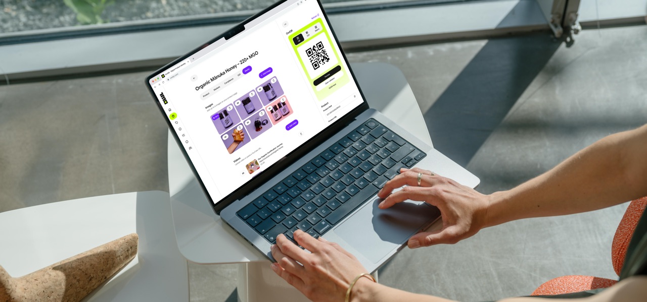 A person works on a laptop at a bright window table. The screen shows an OwlQR product page for organic Manuka honey 220+ MGO, with its GS1 QR code ready to publish.